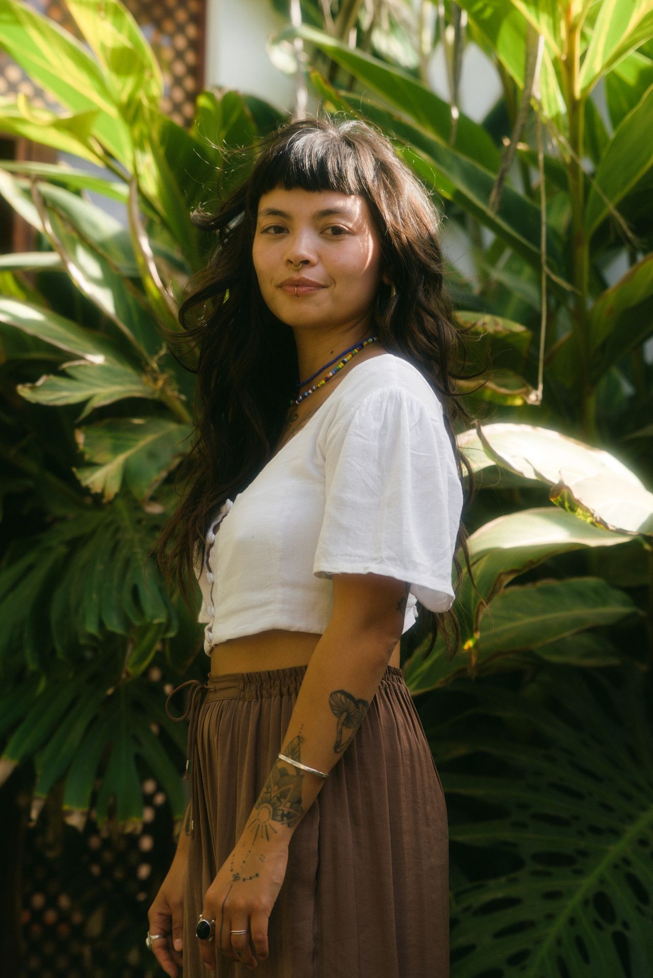 Woman with tattoos wearing white blouse and brown skirt stands in front of lush green plants in sunlight.
