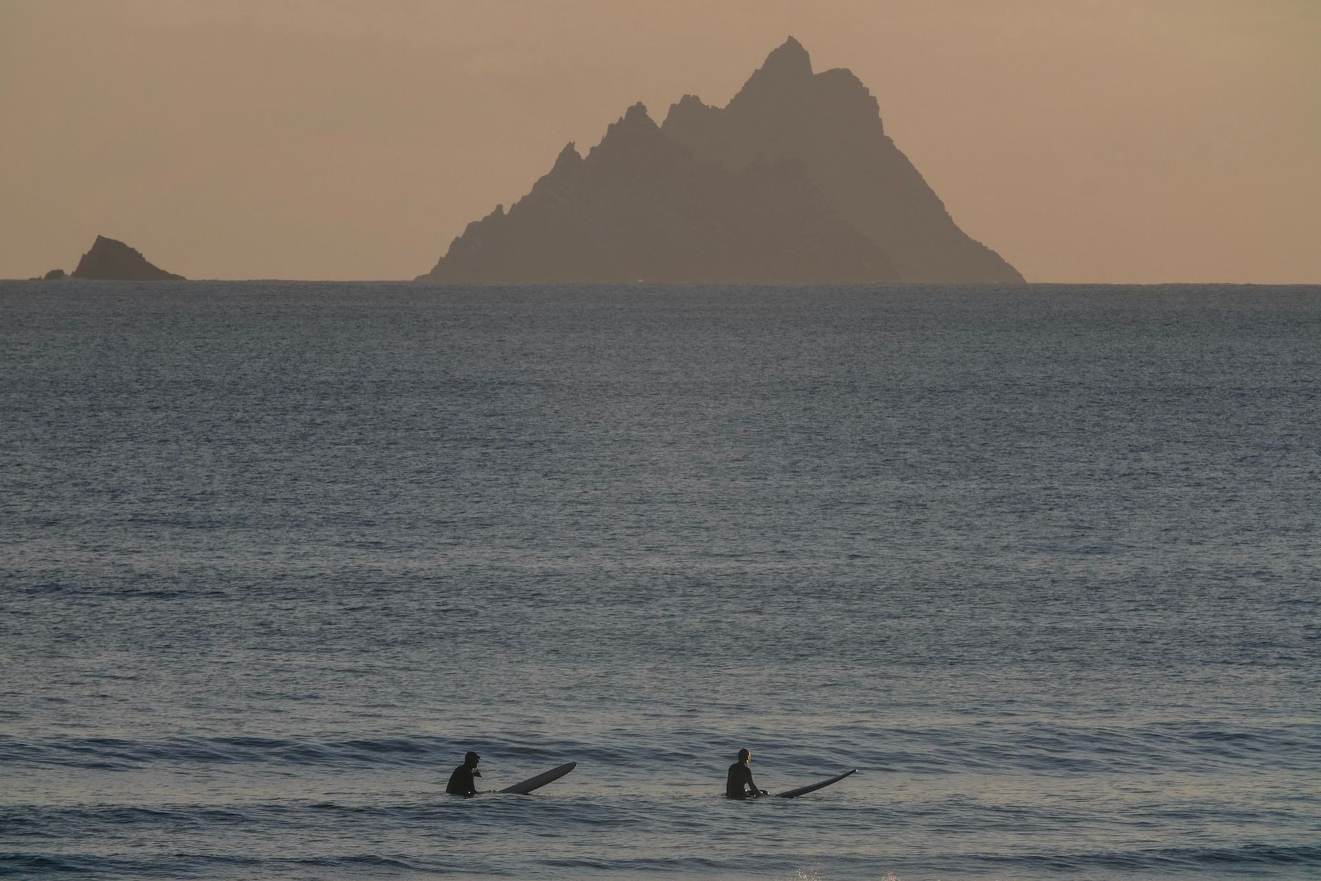 Two surfers waiting in the ocean with distant rocky island at sunset.